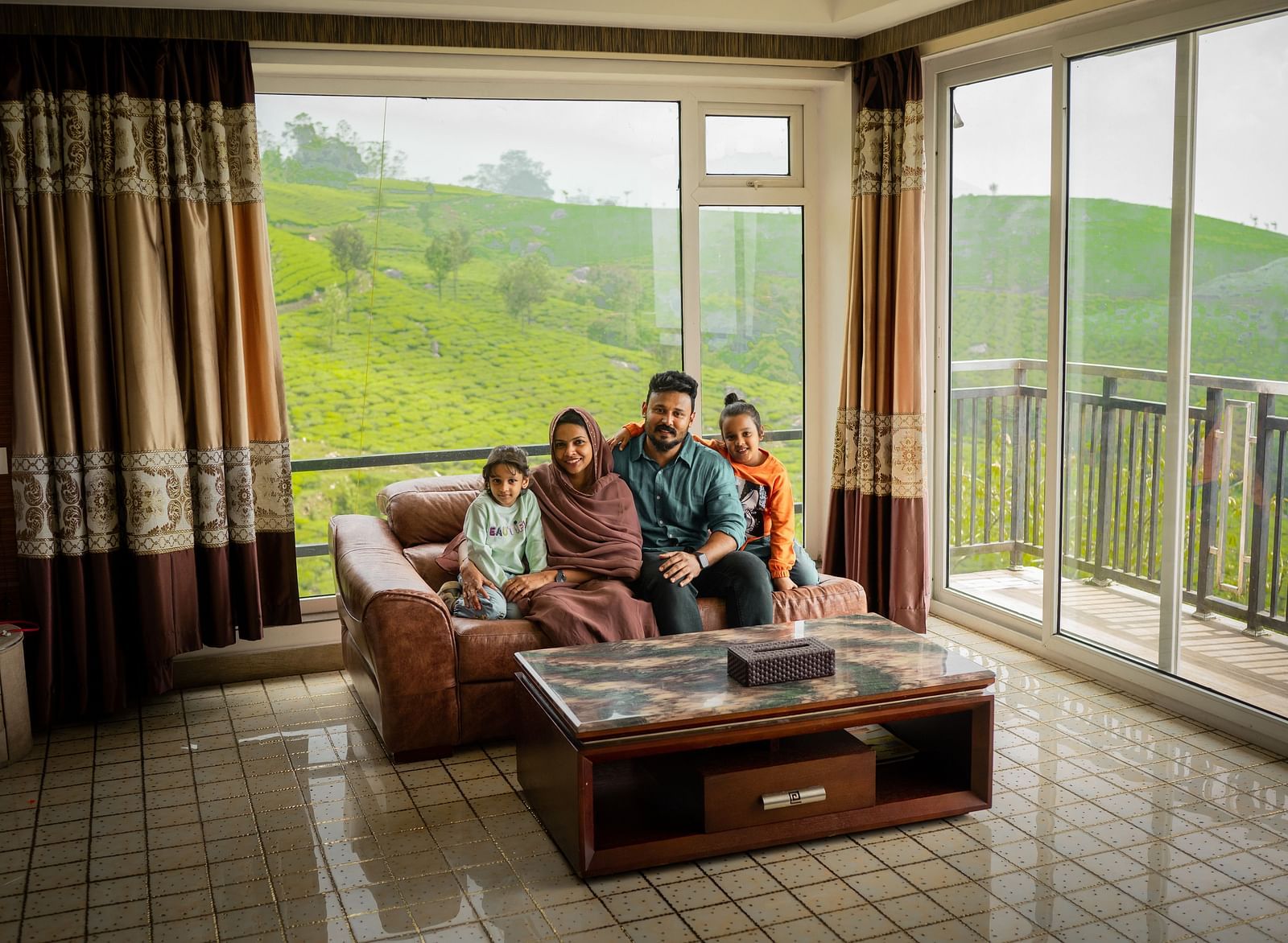 A family of four sitting on a sofa in a room at Parakkat Nature Hotels  and Resorts and outside the balcony there is a view of green hills