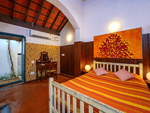 A cosy bedroom featuring a bed with striped orange and yellow linens, wood-panelled walls, an arched ceiling, and an open doorway leading to a small courtyard at Neemrana's Coconut Alley - 21st century, Tranquebar.