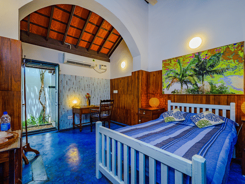 The King Room at Neemrana's Coconut Alley in Tranquebar, featuring a cosy bed with a painting on the wall above the headboard at a distance from the work desk next to the doorway