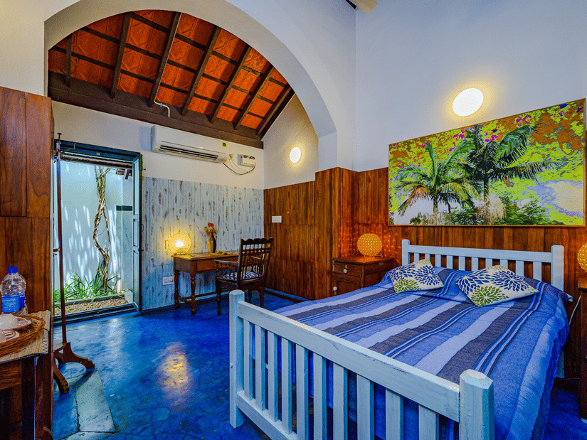 The King Room at Neemrana's Coconut Alley in Tranquebar, featuring a cosy bed with a painting on the wall above the headboard at a distance from the work desk next to the doorway