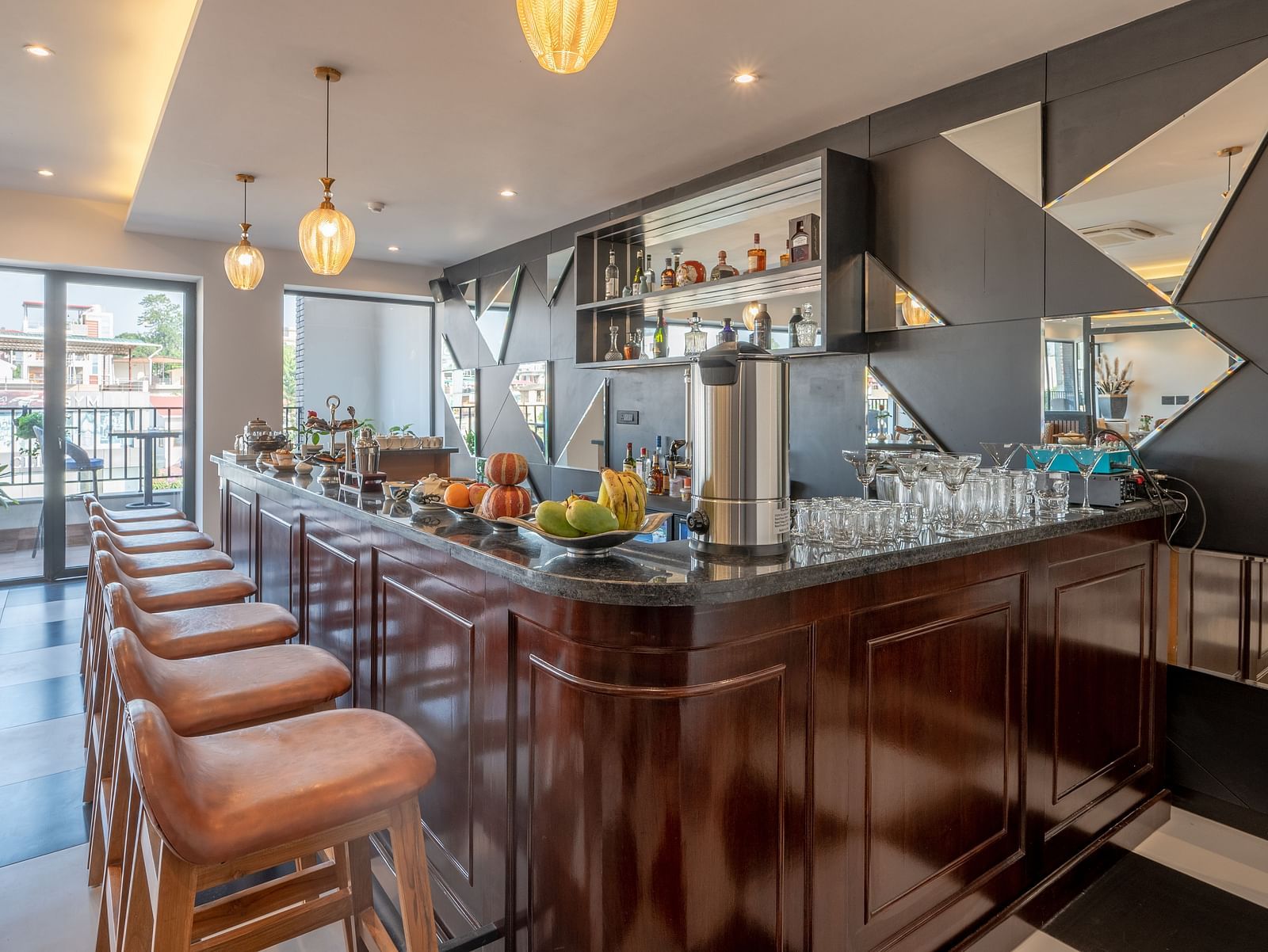 A dimly lit bar and lounge in Dehradun at Vishranti Hotel shows a dark wooden bar counter with stools, a selection of beverages, and a mirrored wall with geometric patterns. 