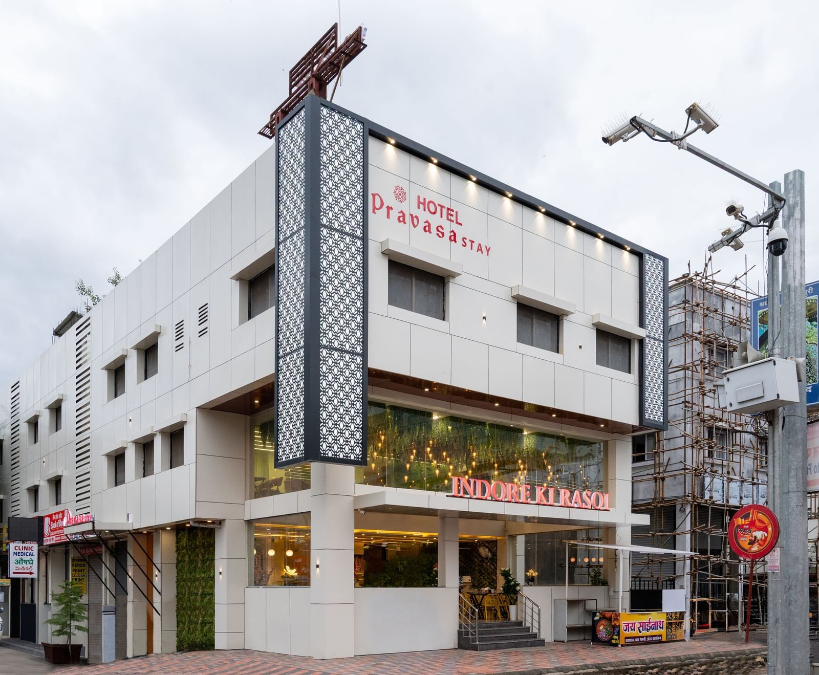 The exterior of Pravasa Stay, Shirdi is a three-storey building with large storefront windows at the ground level.