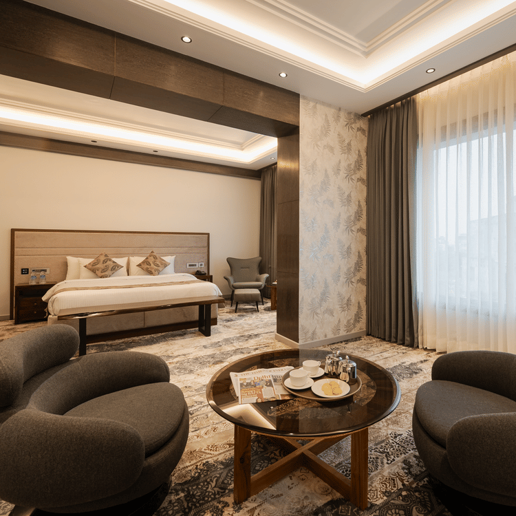 A spacious guest suite with a king-sized bed and two modern grey armchairs around a coffee table at Ananta Express, Katra.
