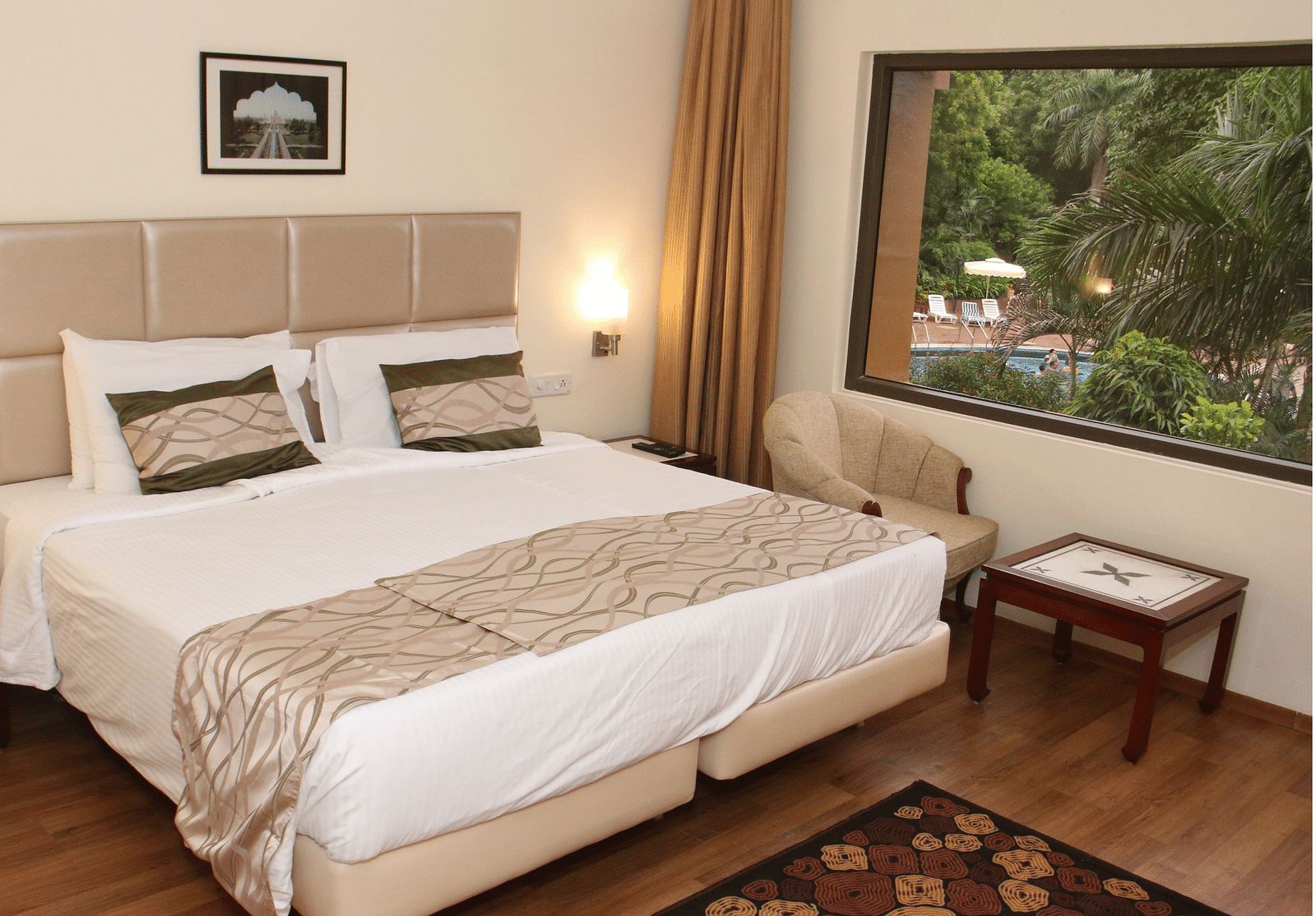 A room with a large bed, beige headboard, wooden flooring, bedside tables, and a window overlooking greenery and a swimming pool - Hotel Clarks Shiraz, Agra
