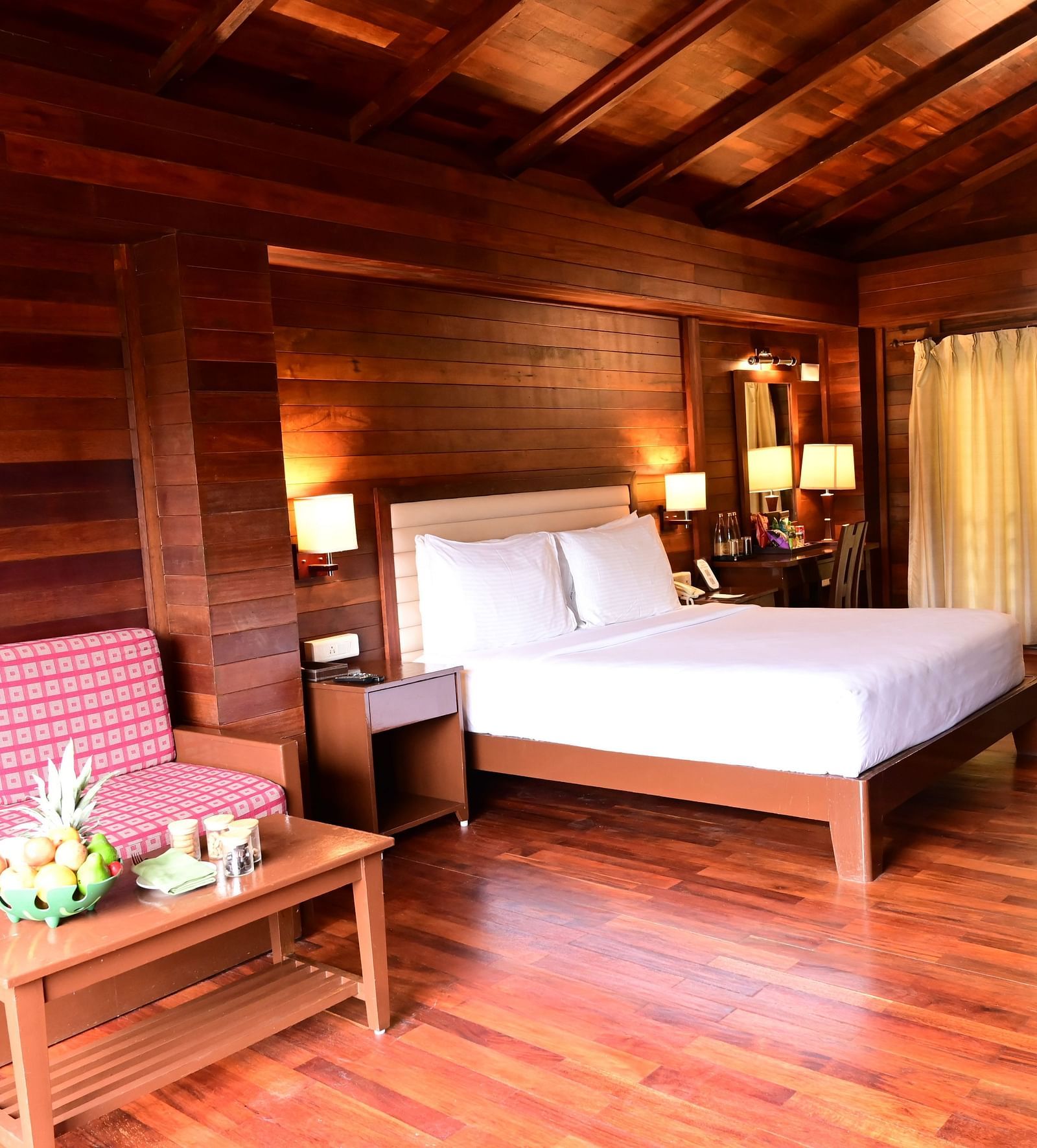 An image of a wood finish room with a king size bed, bed side table, lamps, sofa, table, and decorative items with bright lighting - GReaT trails Kodaikanal by GRT Hotels