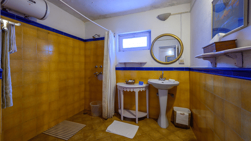 A white & yellow themed bathroom with water heater on the left and shower space beside - Ramgarh Bungalows, Nainital.