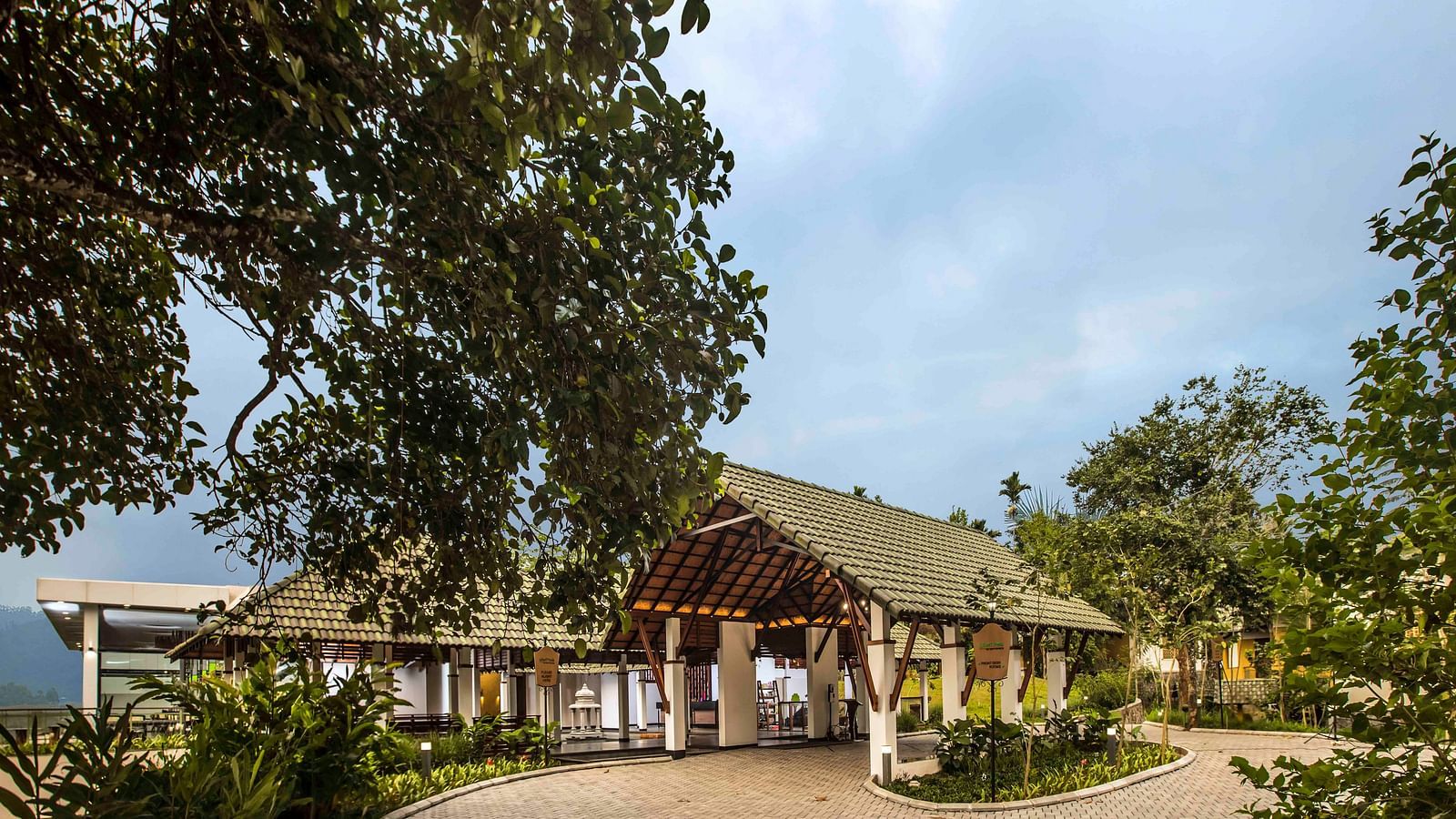 Lush foliage frames a building with a pitched roof and covered walkway - Great Trails Wayanad by GRT hotels.