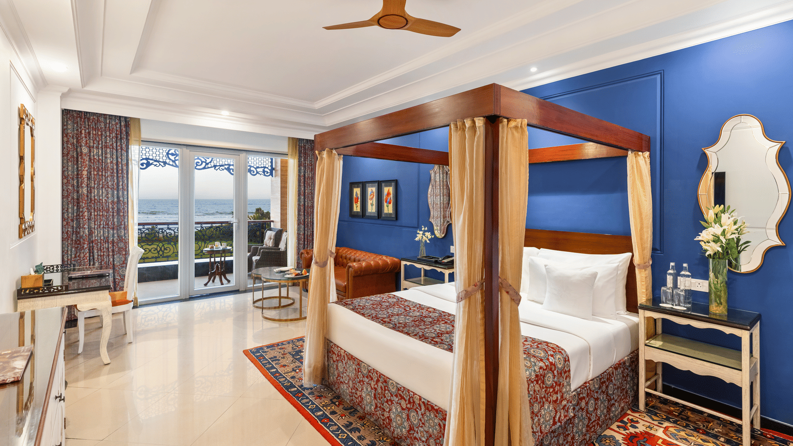Room with a wooden four-poster bed, an armchair, a writing desk and a balcony overlooking the sea at MAYFAIR Palm Beach Resort, Gopalpur-on-Sea.