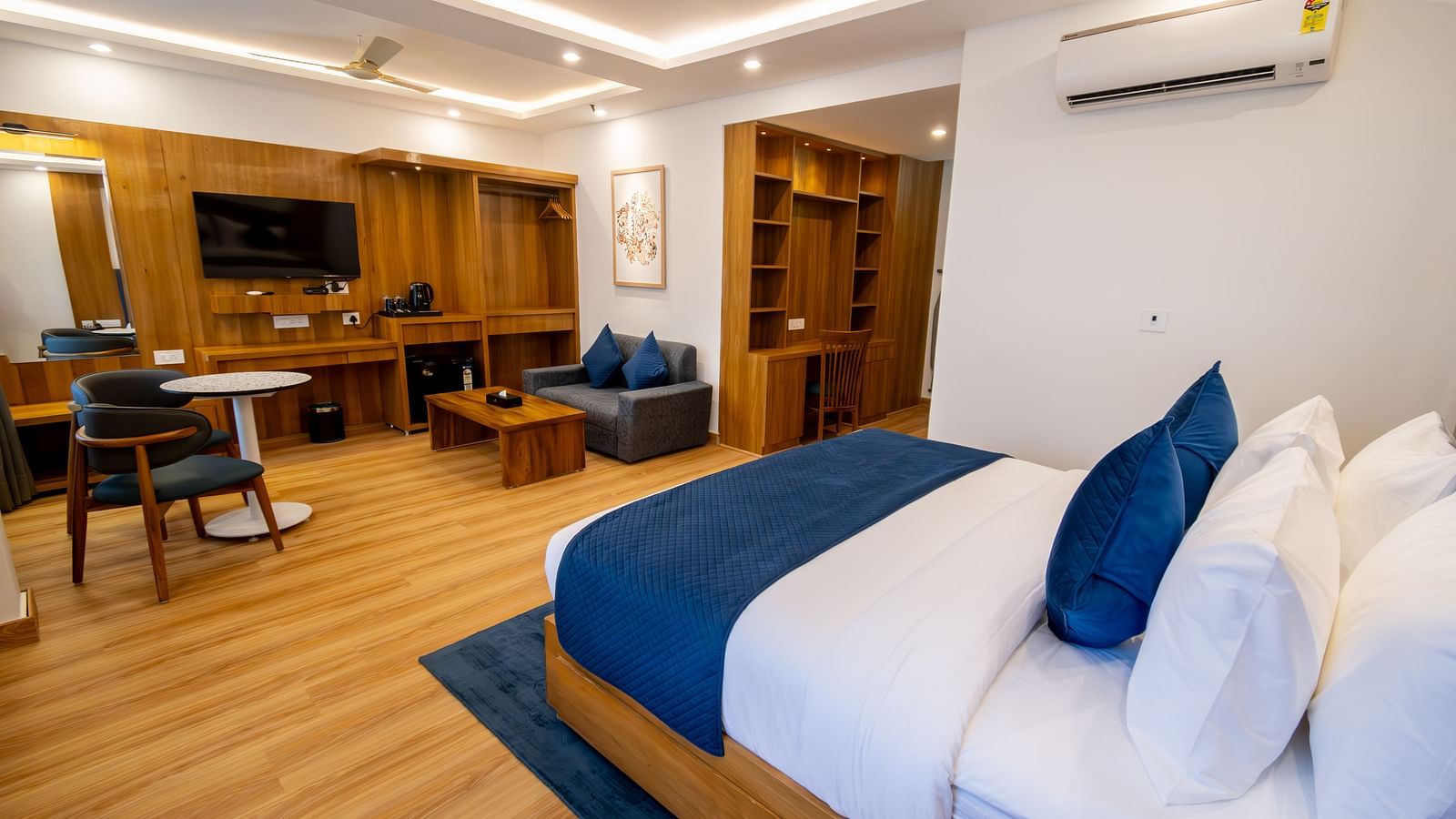 Long shot of a well furnished room with wooden furnishings, king size bed with pillows and blanket, a seating area, flat-screen television, a tea table and a wardrobe | The Tattva Devprayag 