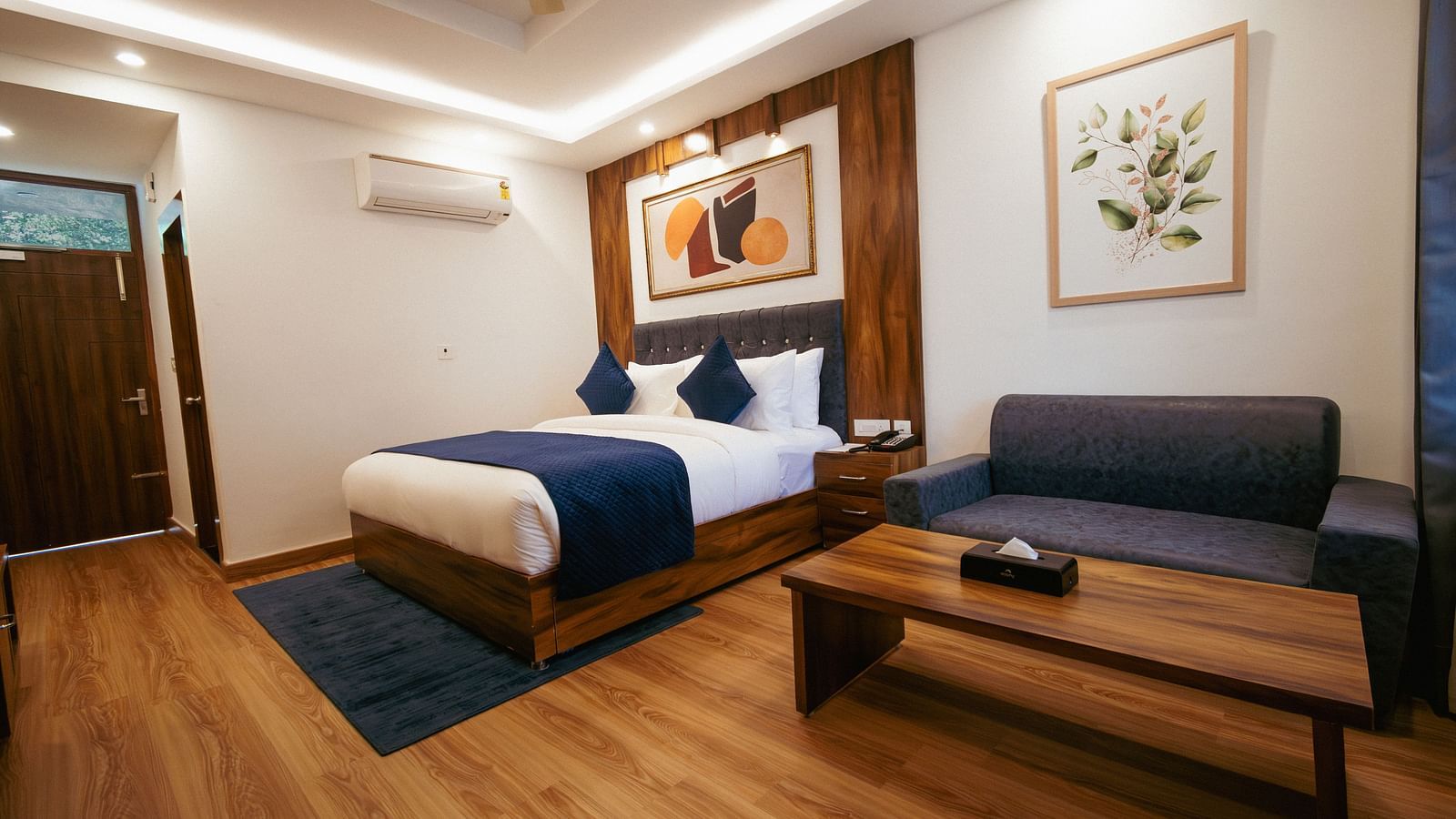 Image of a furnished room with neutral coloured walls featuring a king size bed, a sofa, table and wall paintings | The Tattva Devprayag