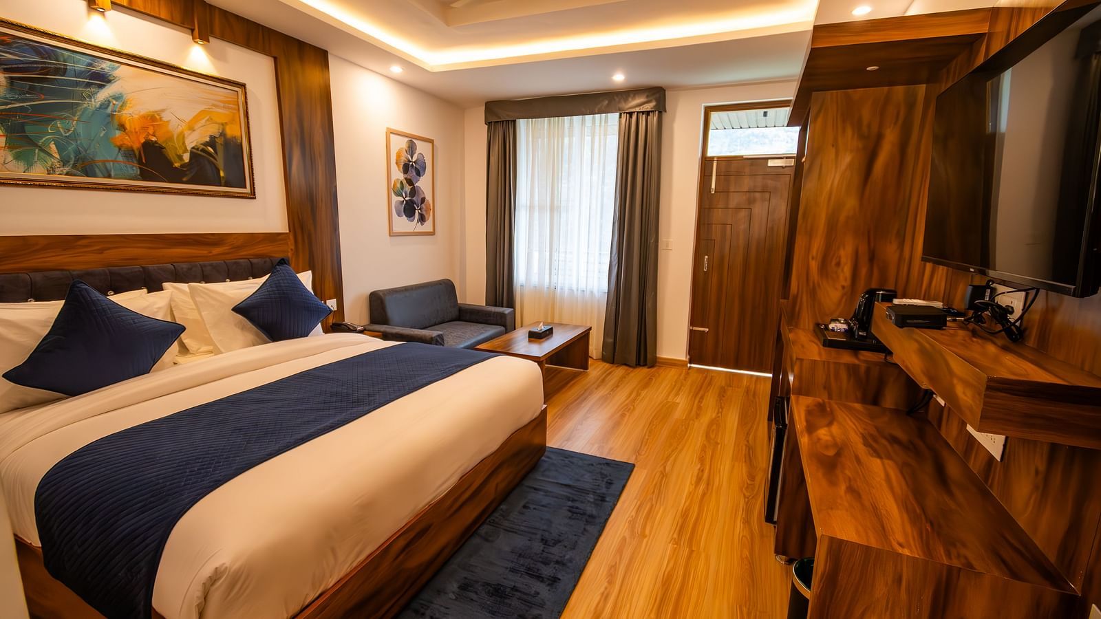 A low angle shot of a well lit, furnished room featuring king size bed, blue pillows, white sheets, wooden flooring and wall paintings along with an expansive window | The Tattva Devprayag