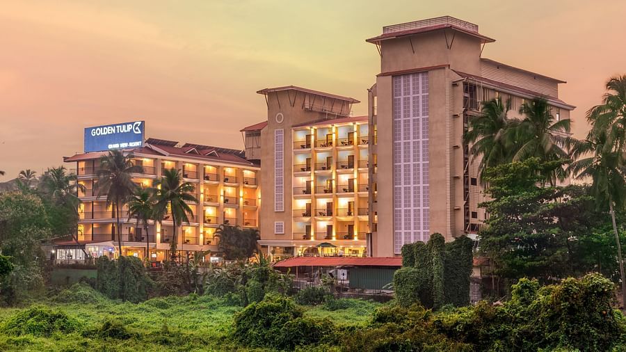 towering hotel buildings featuring balconies, a signage with hotel name, and surrounded by lush greenery, set against a golden hue filled sky - Golden Tulip, Candolim, Goa