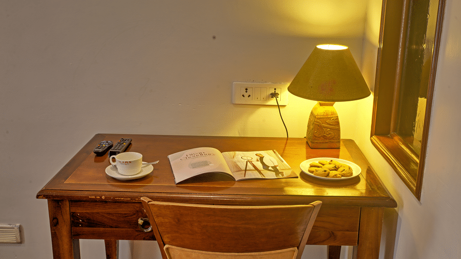 Wooden writing desk with lamp, book, and a cup placed beside a window at Ilara Hotels & Spa, OMR, Chennai