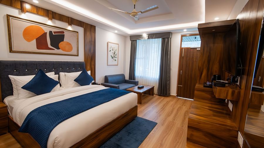 Image of a furnished room with neutral coloured walls featuring a king size bed, a wall painting , blue pillows, TV cabinet and a couch and table | The Tattva Devprayag