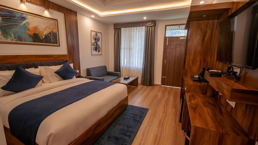 A low angle shot of a  furnished room with dim lighting featuring king size bed, blue pillows, white sheets, wooden flooring and wall paintings along with an expansive window | The Tattva Devprayag