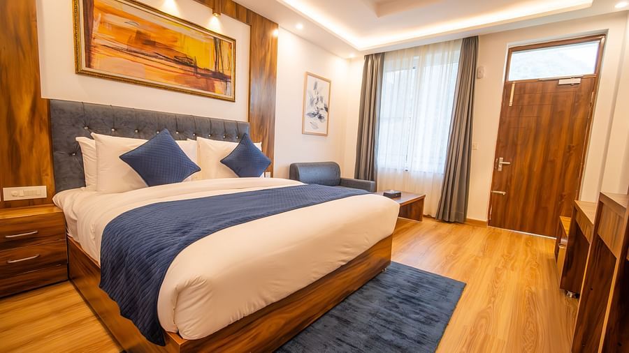 A low angle shot of a well lit, furnished room featuring king size bed, blue pillows, white sheets, wooden flooring and wall paintings along with an expansive window | The Tattva Devprayag