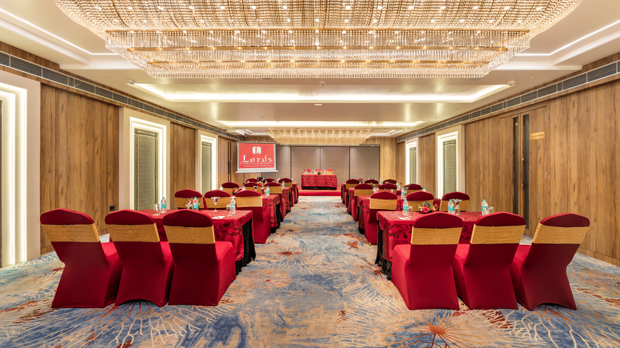Spacious banquet hall at Lords Inn Sangam, Bhopal set up in theatre style with rows of red chairs, patterned carpet, and elegant ceiling lighting.