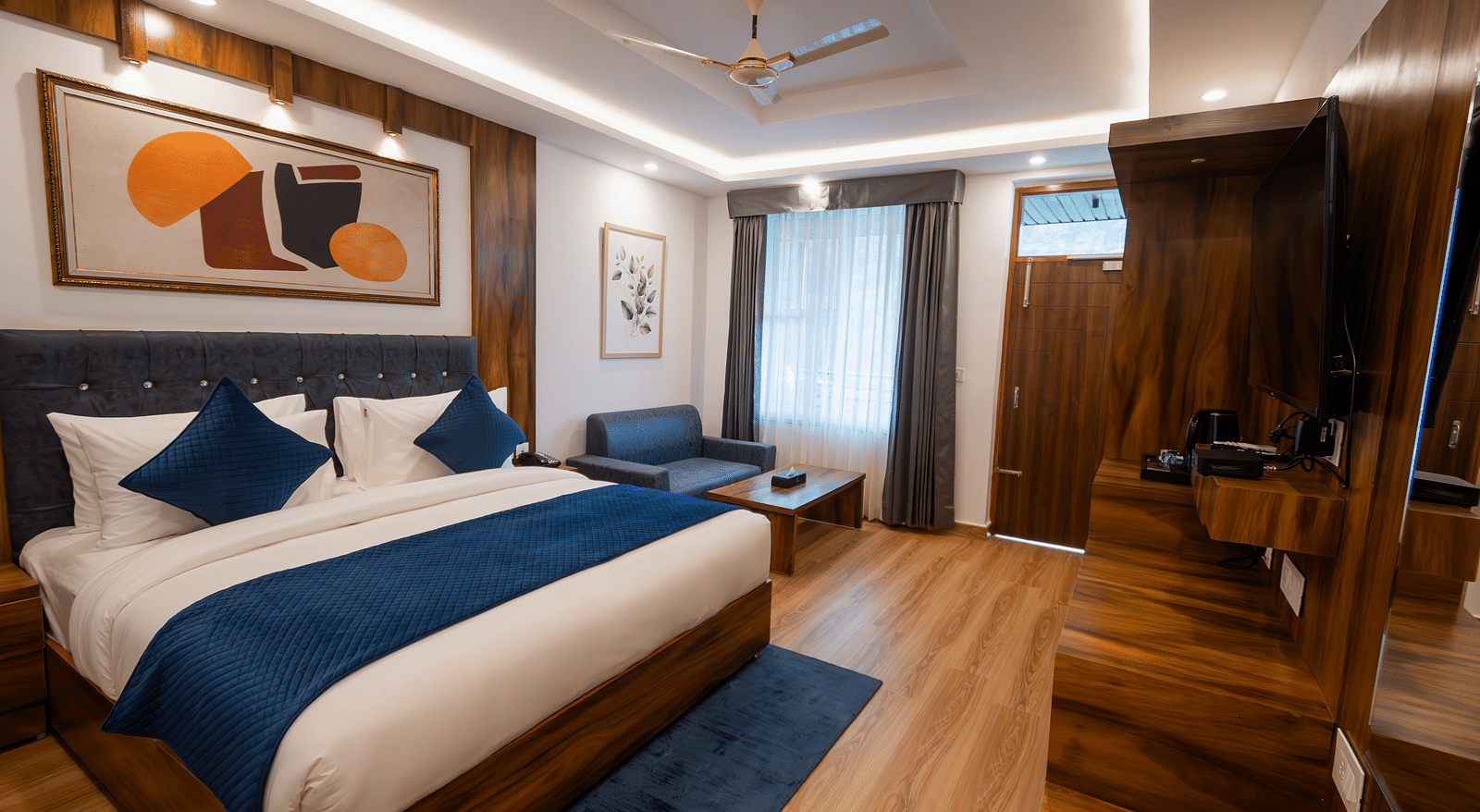 Image of a furnished room with neutral coloured walls featuring a king size bed, a wall painting , blue pillows, TV cabinet and a couch and table | The Tattva Devprayag