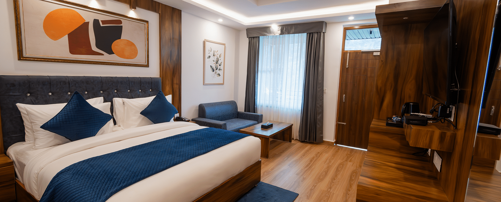 Image of a furnished room with neutral coloured walls featuring a king size bed, a wall painting , blue pillows, TV cabinet and a couch and table | The Tattva Devprayag