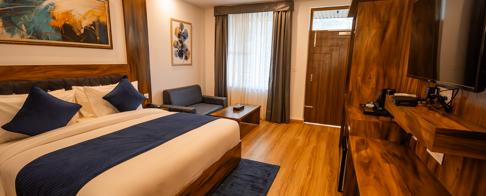 A low angle shot of a well lit, furnished room featuring king size bed, blue pillows, white sheets, wooden flooring and wall paintings along with an expansive window | The Tattva Devprayag