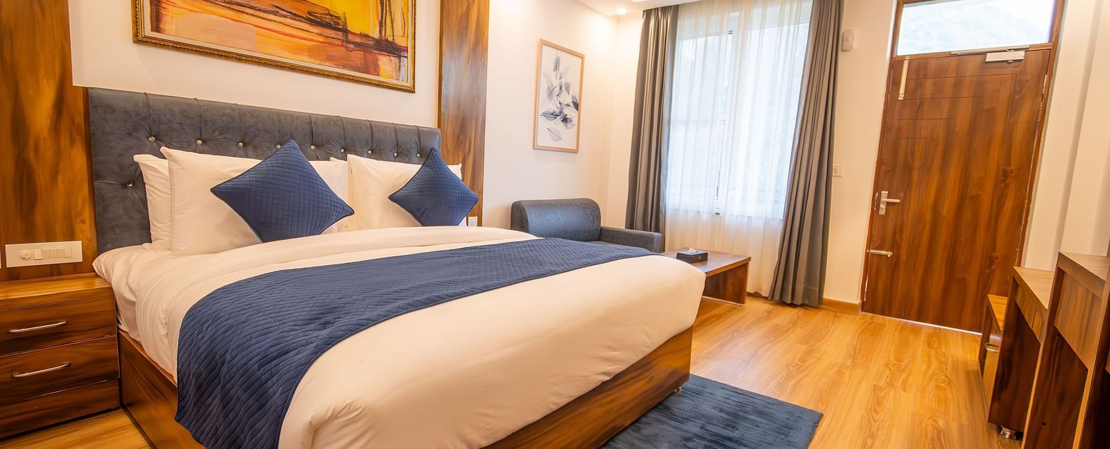 A low angle shot of a well lit, furnished room featuring king size bed, blue pillows, white sheets, wooden flooring and wall paintings along with an expansive window | The Tattva Devprayag