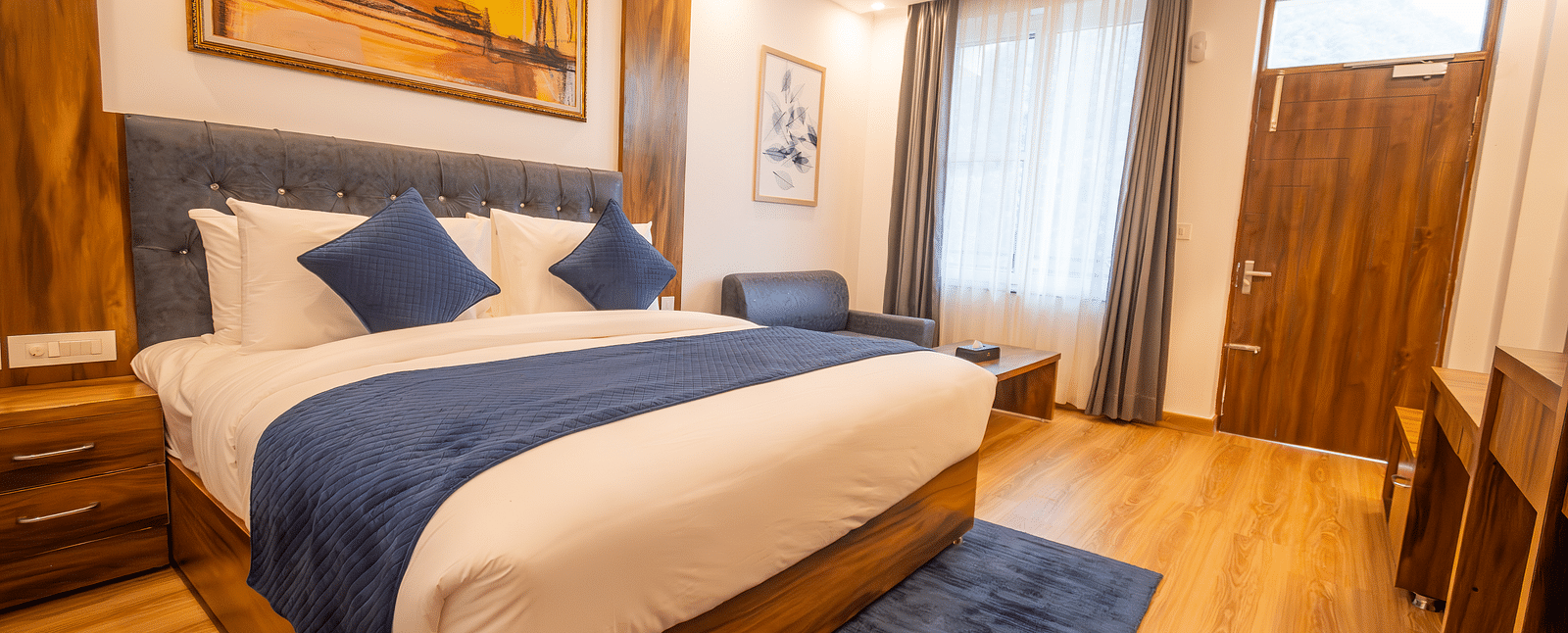 A low angle shot of a well lit, furnished room featuring king size bed, blue pillows, white sheets, wooden flooring and wall paintings along with an expansive window | The Tattva Devprayag