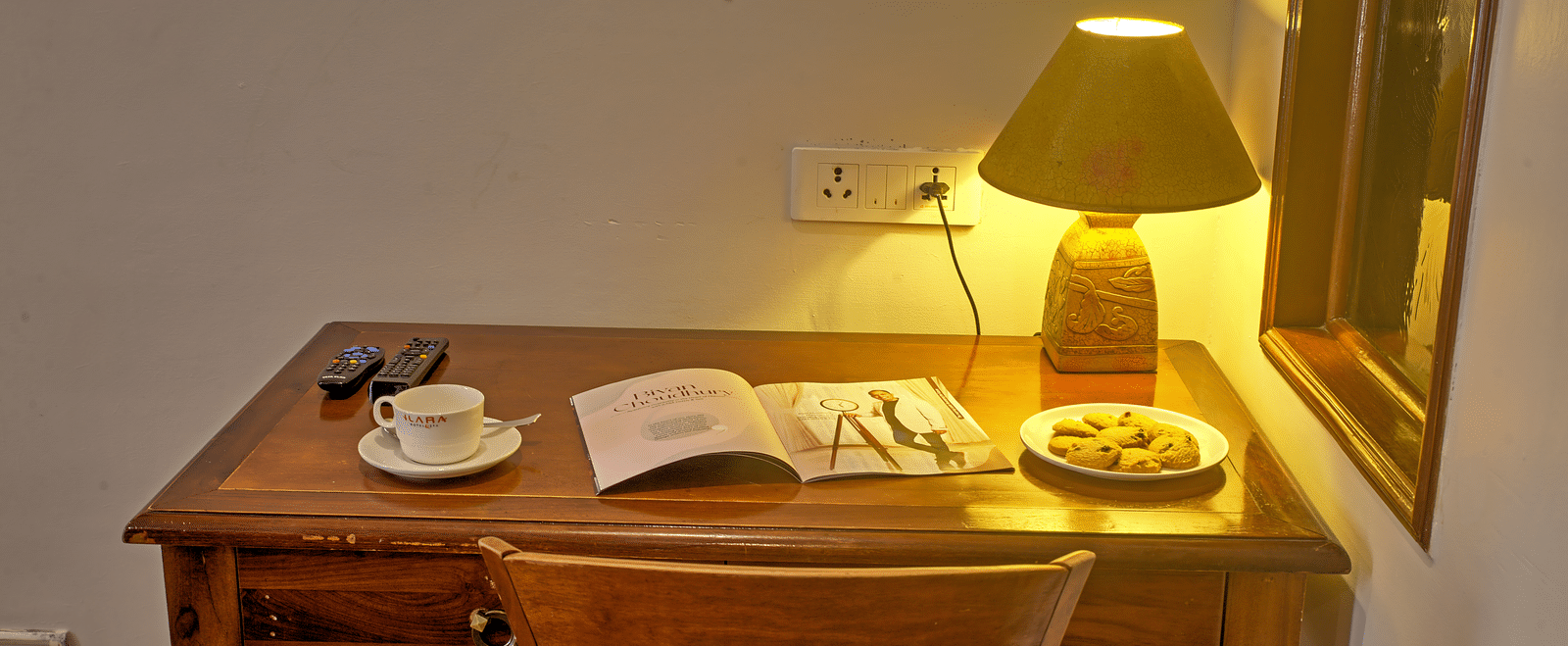 Wooden writing desk with lamp, book, and a cup placed beside a window at Ilara Hotels & Spa, OMR, Chennai
