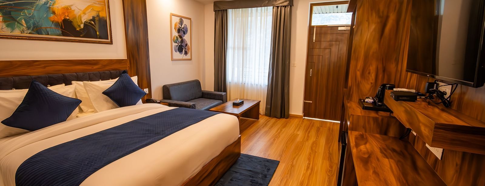 A low angle shot of a well lit, furnished room featuring king size bed, blue pillows, white sheets, wooden flooring and wall paintings along with an expansive window | The Tattva Devprayag