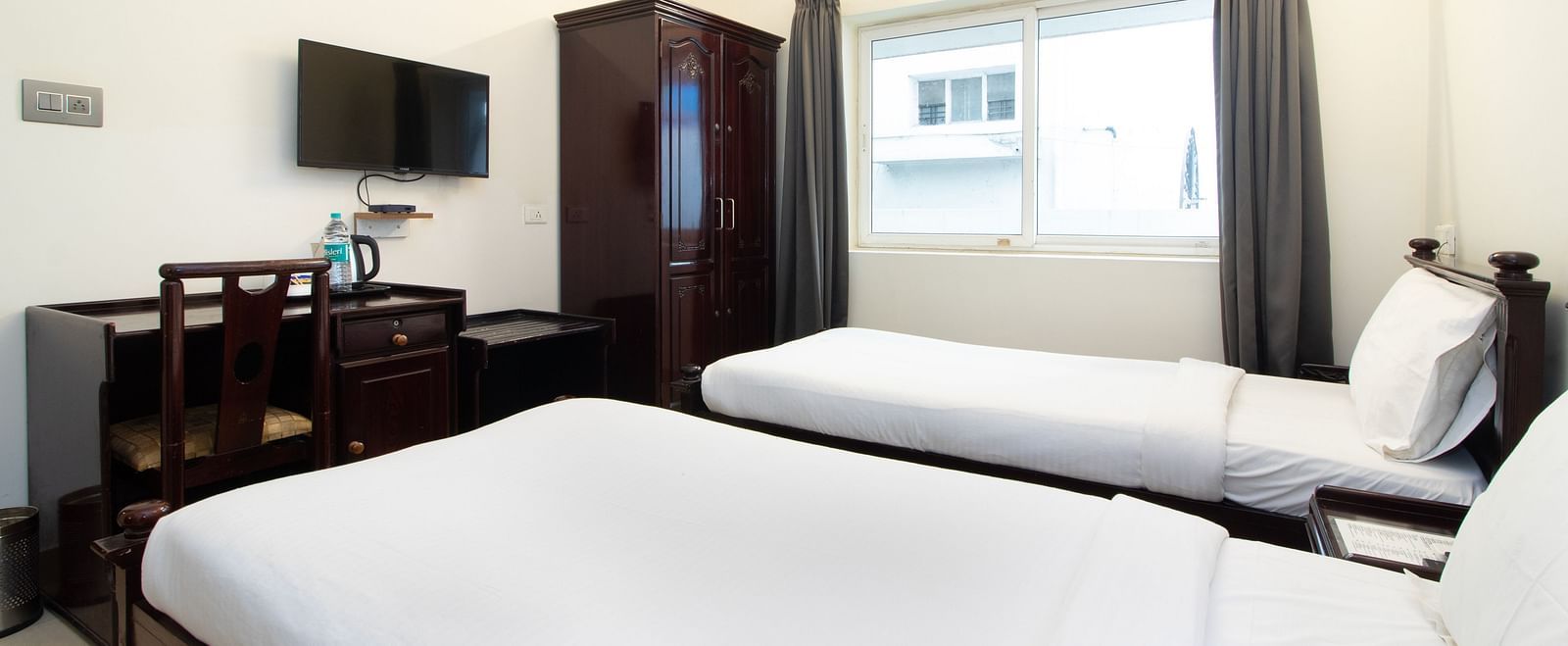 A well-lit room showing a double bed with white bedding, a chair, a desk area with a television, and a window for natural light at Sanctum Suites Indiranagar, Bangalore.