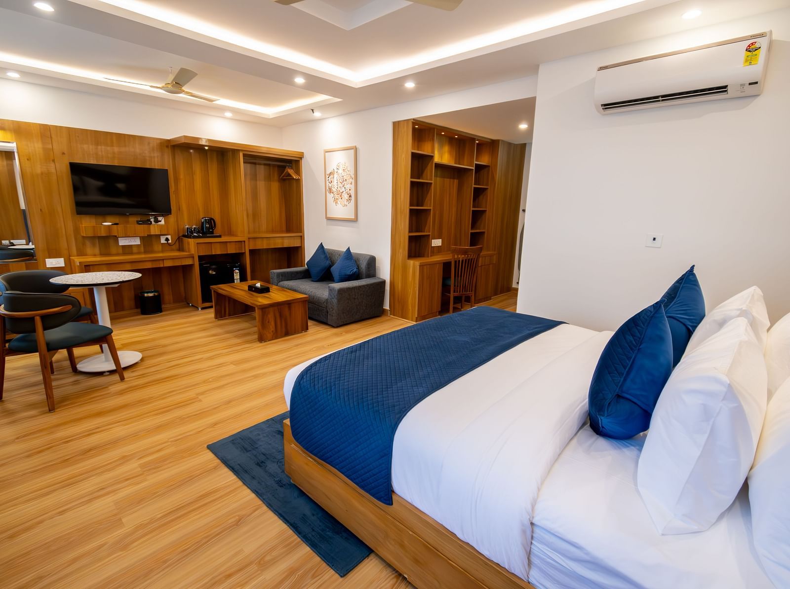 Long shot of a well furnished room with wooden furnishings, king size bed with pillows and blanket, a seating area, flat-screen television, a tea table and a wardrobe | The Tattva Devprayag 