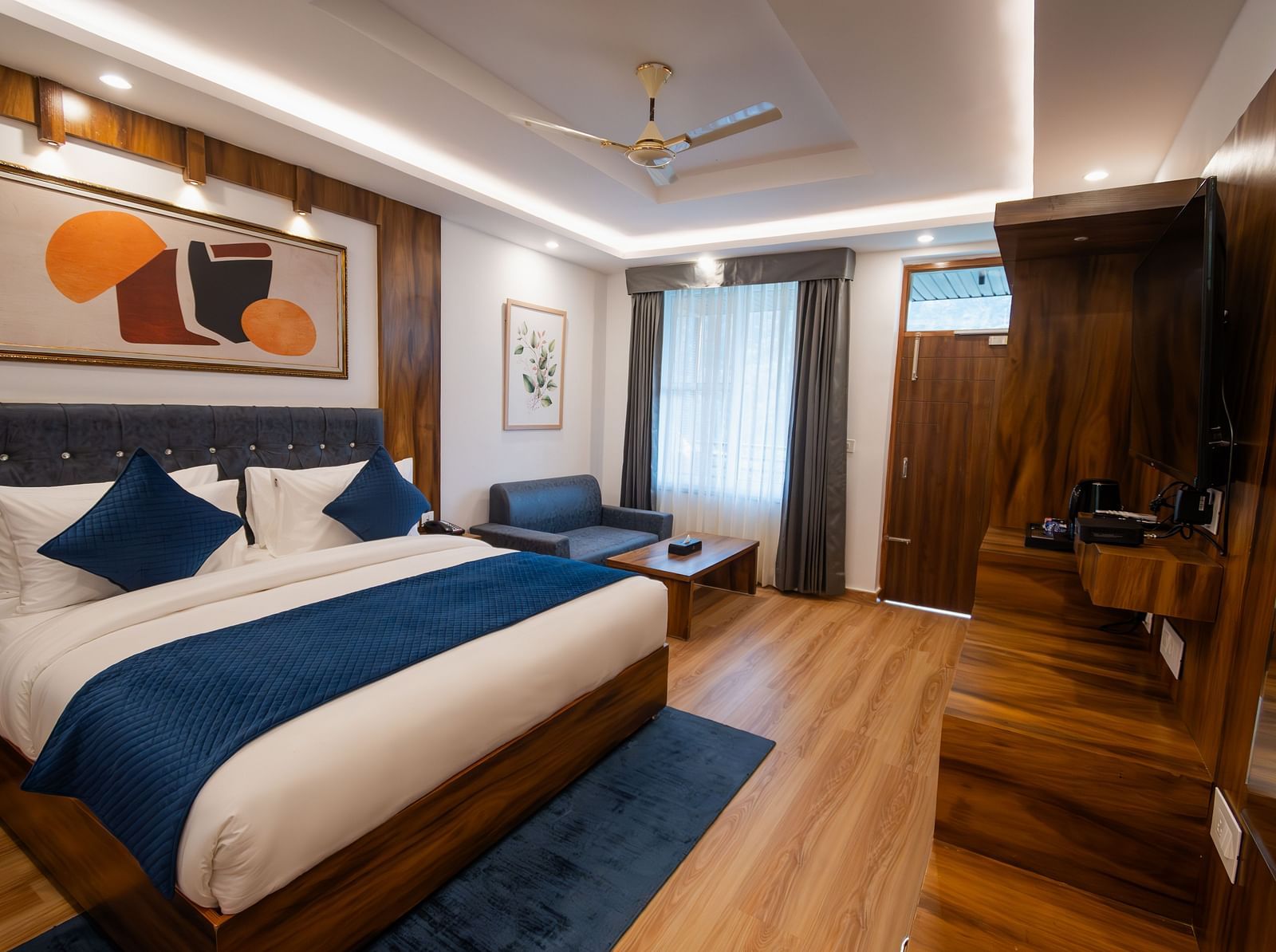 Image of a furnished room with neutral coloured walls featuring a king size bed, a wall painting , blue pillows, TV cabinet and a couch and table | The Tattva Devprayag