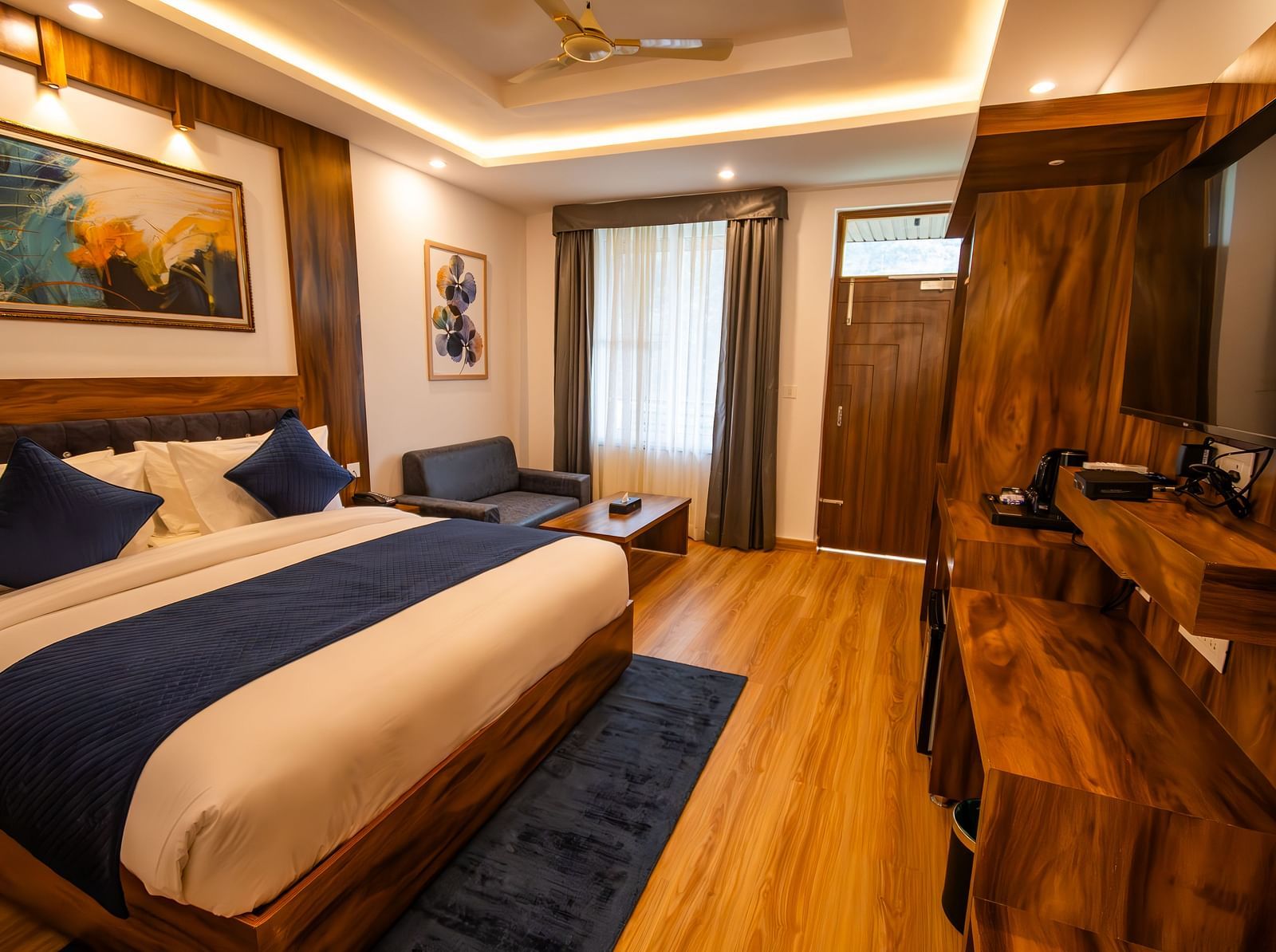 A low angle shot of a well lit, furnished room featuring king size bed, blue pillows, white sheets, wooden flooring and wall paintings along with an expansive window | The Tattva Devprayag