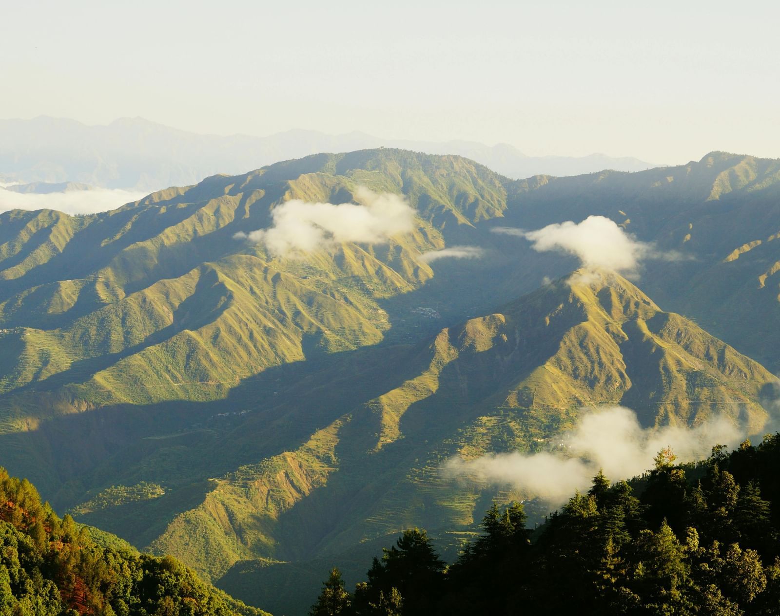 Sunlight casts long shadows over a range of forested mountains with scattered clouds drifting across the ridges under a clear sky.