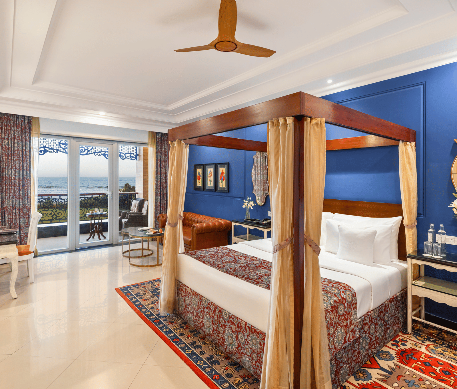 Room with a wooden four-poster bed, an armchair, a writing desk and a balcony overlooking the sea at MAYFAIR Palm Beach Resort, Gopalpur-on-Sea.