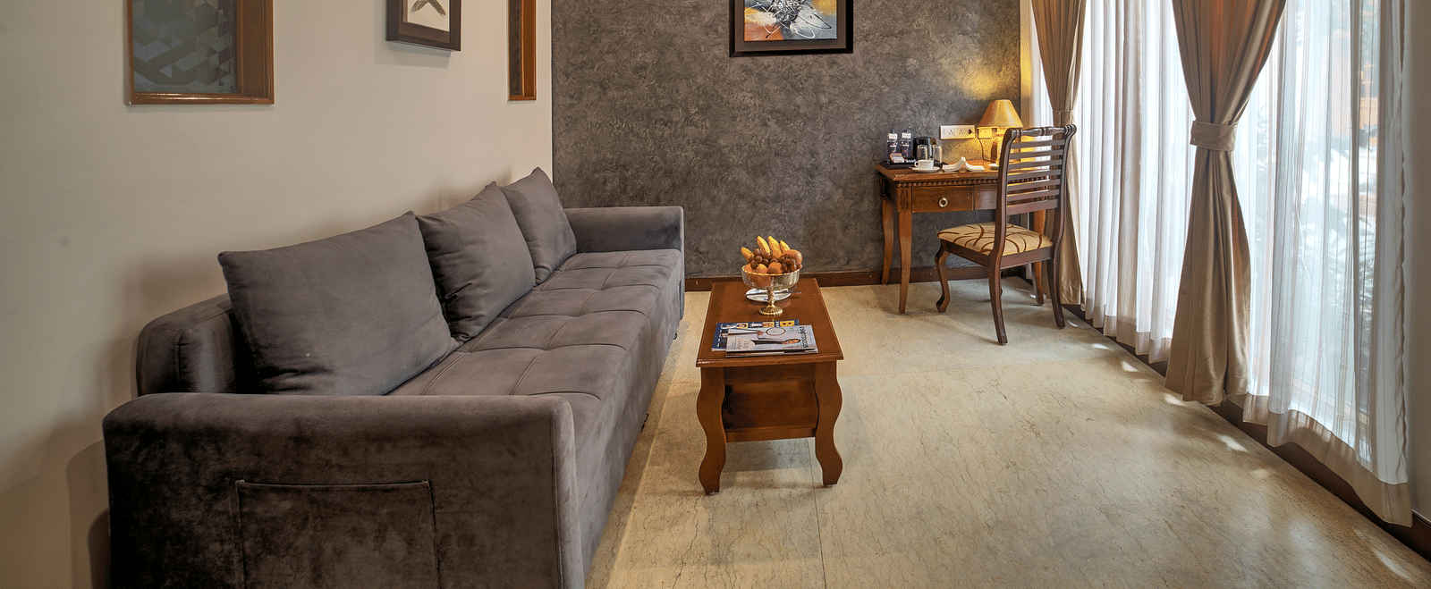 Spacious suite living room with plush grey sofa and natural light from full-length curtains at Ilara Hotels & Spa, OMR, Chennai