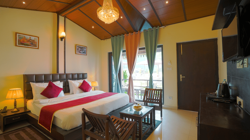 A neatly arranged room with a big bed, 2 wooden chairs on either side of a coffee table, a chandelier and full length curtains | Sol Glow Resort, Nainital