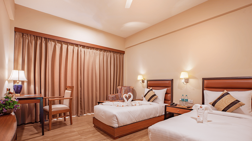 A spacious bedroom at The Lalita Grand Mathura - Vrindavan featuring twin beds, decorative towel art, wooden furniture, warm lighting, and long curtains.