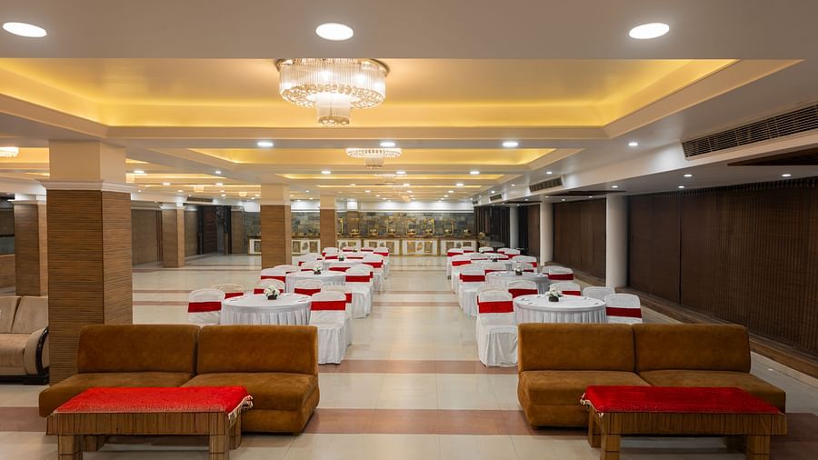 A banquet hall with rows of round tables and white chairs, featuring a seating area with plush brown sofas accented with red cushions. Chandeliers and recessed ceiling lights provide a warm ambience. - Clarks Inn Express, Near Jammu Bari Brahmana Railway S