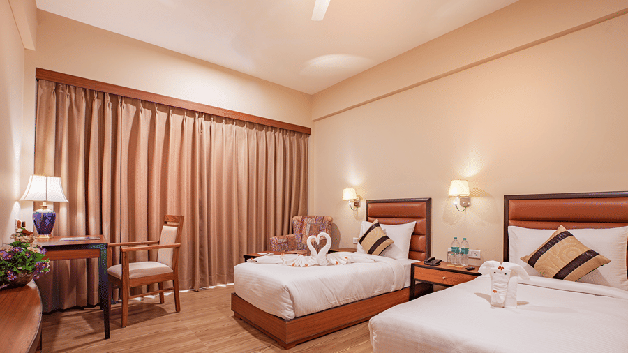 A spacious bedroom at The Lalita Grand Mathura - Vrindavan featuring twin beds, decorative towel art, wooden furniture, warm lighting, and long curtains.