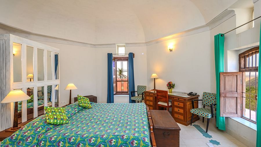 Room with bed, patterned bedding and pillows, furniture including desk, chairs, and trunk, windows with wooden shutters, curtains, decorative wall lights, and unique floor pattern - Tijara Fort-Palace - 19th Century, Alwar.
