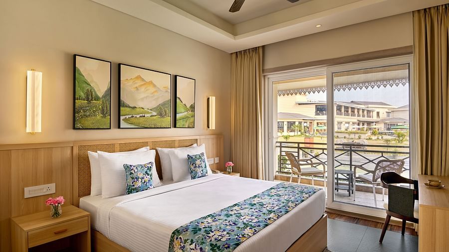 A hotel bedroom with a king bed dressed in white and a blue floral runner. Three landscape paintings hang above the wooden headboard. A nightstand with flowers sits beside the bed. Sliding glass doors with light curtains open to a balcony - Voyage The Soli