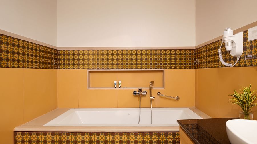 Stylish bathroom featuring a built-in bathtub with patterned yellow and black tile accents, wall-mounted faucet and handheld shower, small shelf niche, and part of a vanity with a vessel sink and countertop - Voyage The Solitaire Bliss.