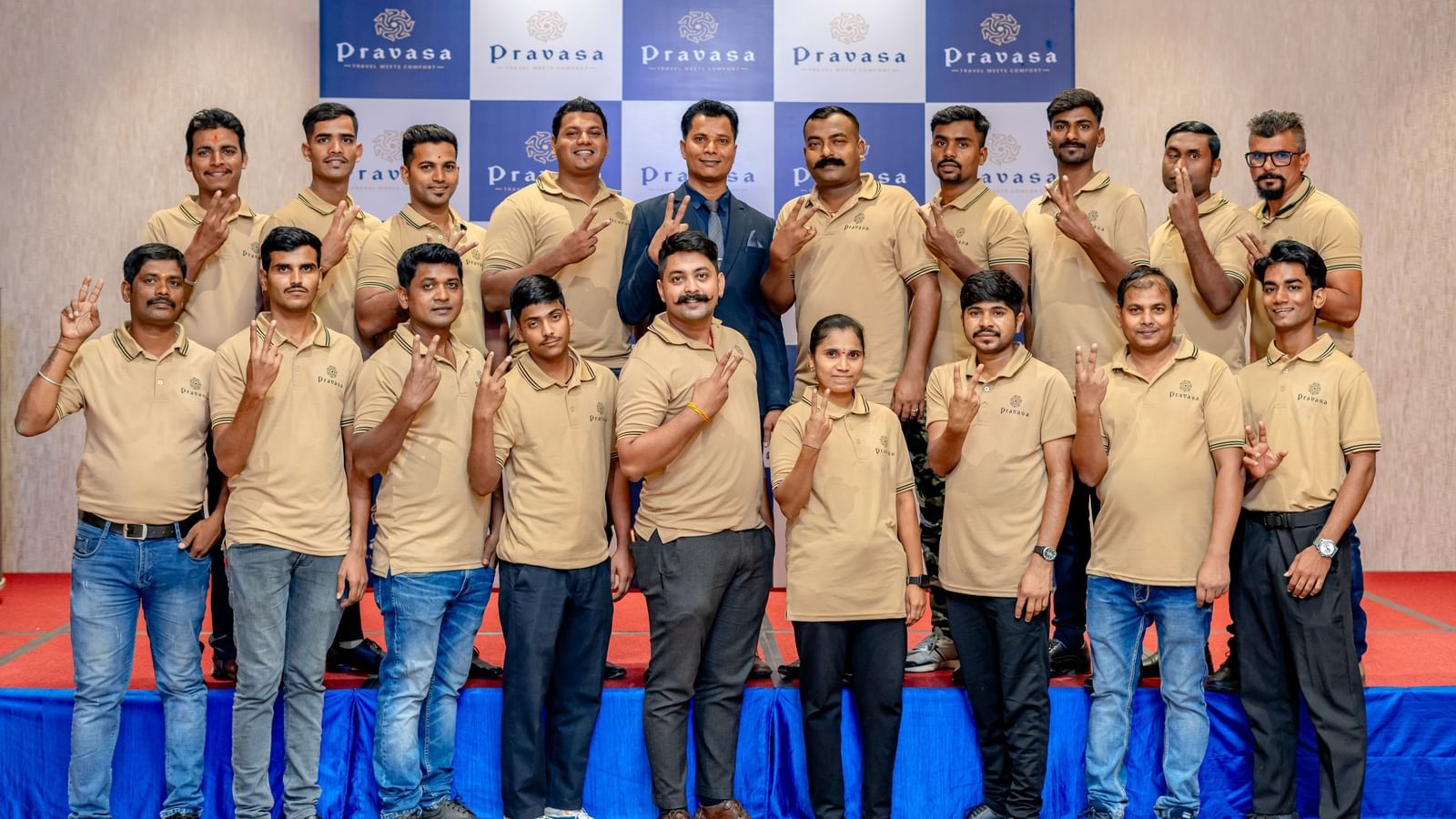 A large group of men in matching tan shirts and blue jeans stand in rows looking at the camera in front of a blue and white backdrop.