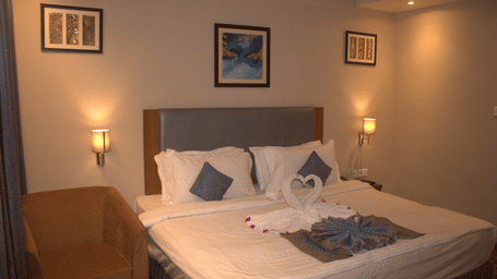 Bedroom with a king size bed, white and blue cushions, framed artwork, bedside lamps, and wooden flooring Pipul Padmaja Premium Hotel & Convention.