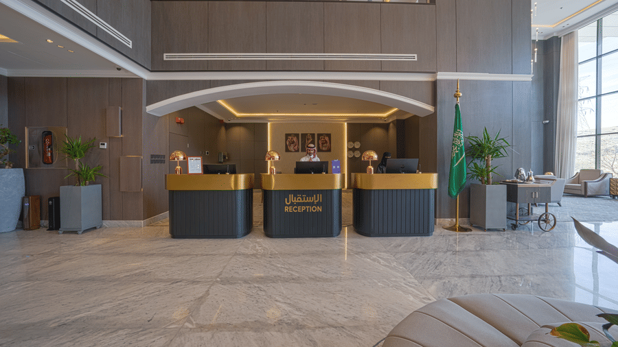 The reception desk of Grand Iva Hotel, Abha, seen with a person looking down into the podium, with natural lighting entering through the glass windows lights the lobby up.