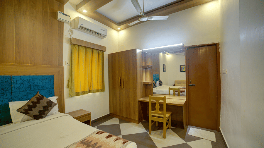 The corner of the Green Room featuring a comfortable bed, vibrant yellow curtains, a wooden wardrobe, and a chequered floor - Kailash Conventions ECR