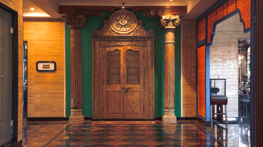 A grand corner hallway with polished flooring, carved wooden double doors, tall columns, wall decor and warm lighting creating a traditional welcome area at MAYFAIR Bay Resort, Paradeep.
