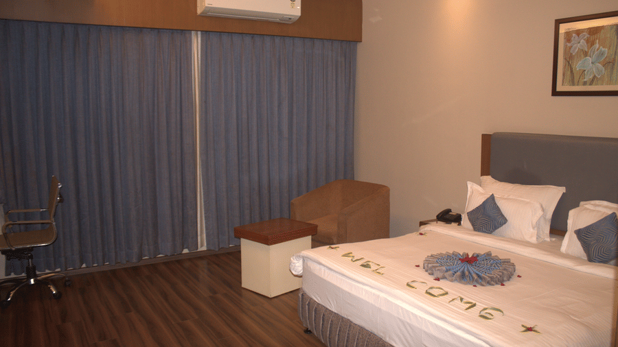 Bedroom with a king size bed, white bedding with a towel design spelling 'Welcome', work desk, office chair, and wide window covered with blue curtains at Pipul Padmaja Premium Hotel & Convention.