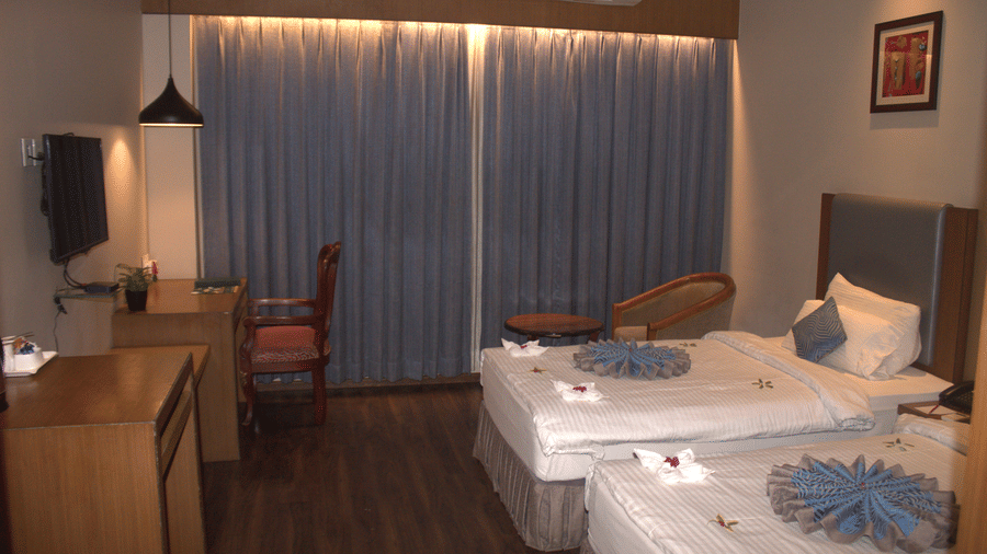 Side view of a room with twin beds, desk and chair on the left, full-length grey blackout curtains, and a mounted light fixture at Pipul Padmaja Premium Hotel & Convention.