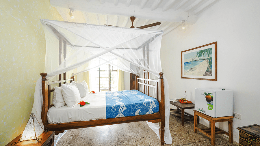 Interior view of a bedroom with a wooden four-poster bed with a mosquito net, a patterned bedspread, a framed picture on the wall, a small refrigerator, and a wooden table at shooting star Boutique Hotel.